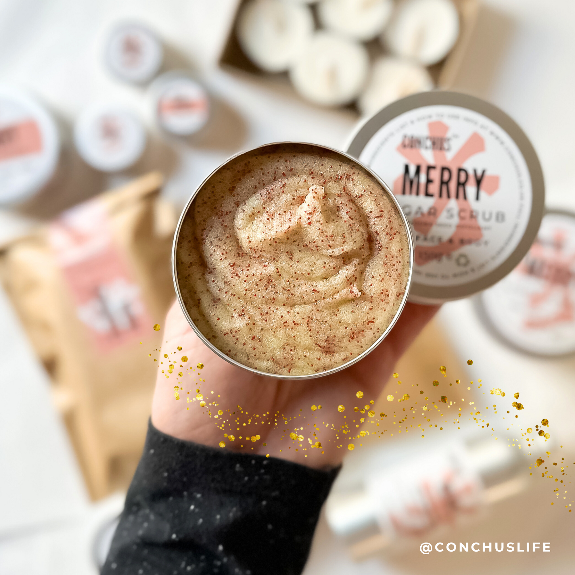 Open tin of CONCHUS MERRY Sugar Scrub held above CONCHUS festive packaging and skincare products