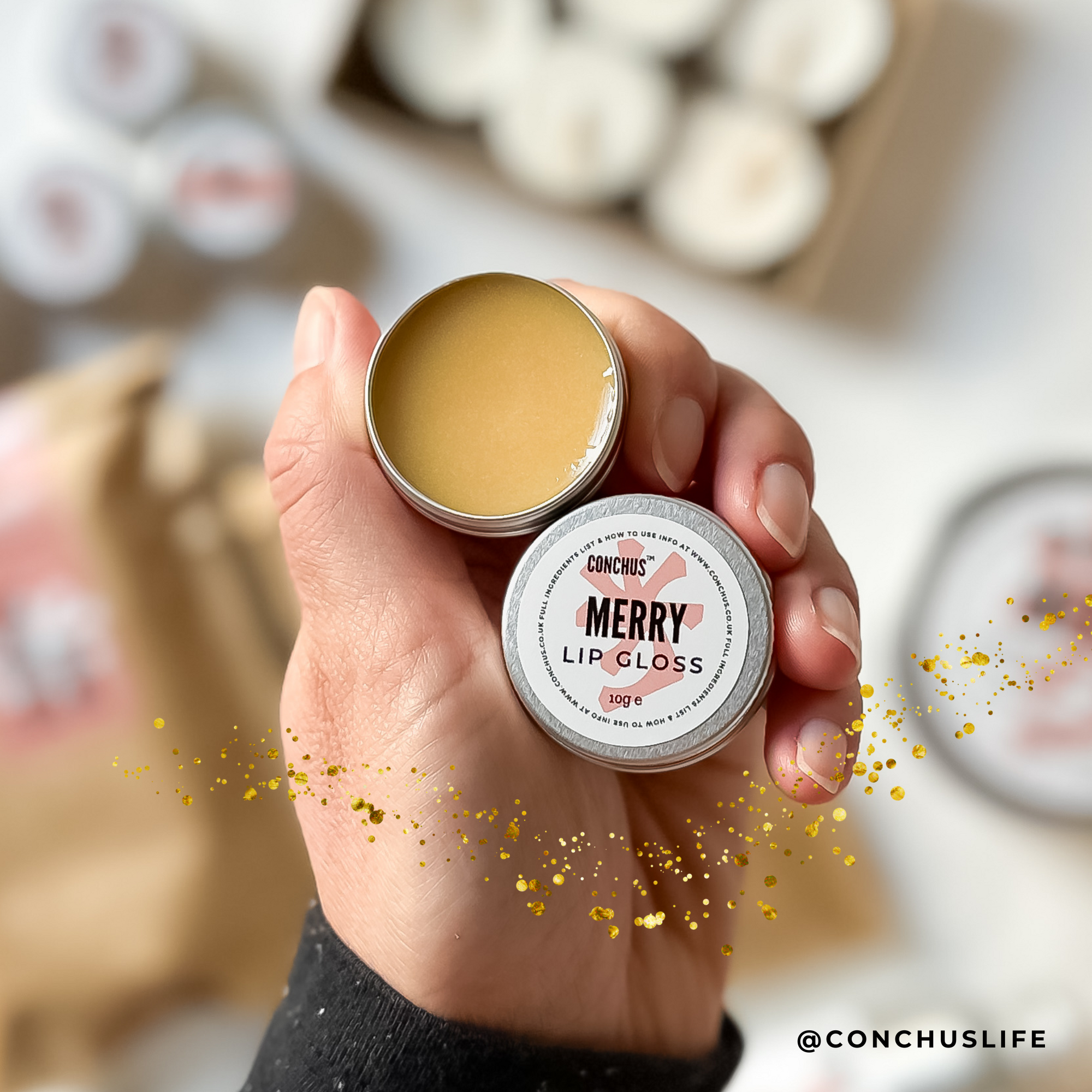 Open tin of CONCHUS MERRY Lip Gloss held above CONCHUS festive skincare packaging