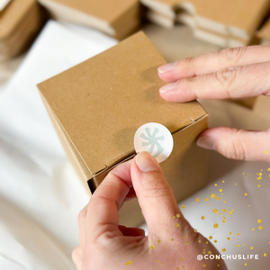 Person sealing kraft gift box for CONCHUS MERRY Botanical Candle with branded sticker
